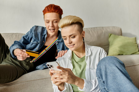 Two short-haired women sit on a couch, engrossed in a cell phones screen, sharing a moment of intimacy and connection.の写真素材