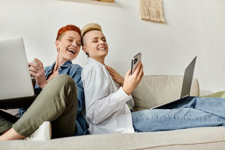 Two lgbt women with short hair studying on laptops together on a cozy couch at home.の写真素材