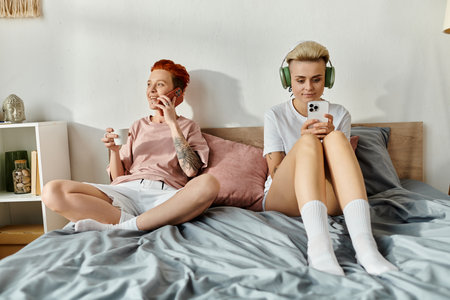 A lesbian couple with short hair enjoying music on headphones together in a cozy bedroom setting.の写真素材