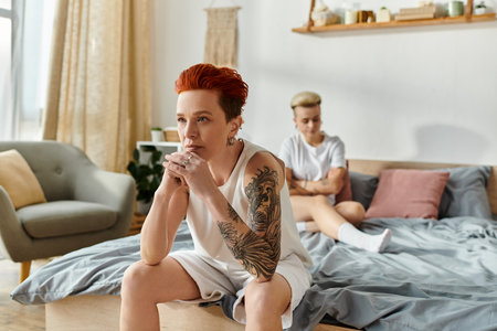 A lesbian couple with short hair sitting on a bed, showcasing their tattoos, having conflictの写真素材