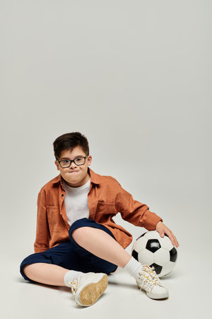 little boy with with Down syndrome with glasses sits on ground with soccer ball.の写真素材