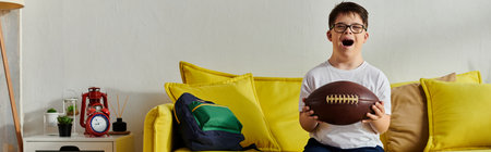 Little child with Down syndrome happily holds a football while sitting on a couch at home.の写真素材
