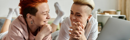 Two women with short hair laying on a bed, engrossed in a laptop screen, sharing a tender moment in...