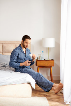 Handsome man peacefully sits on bed, sipping coffee in morning light.