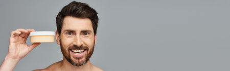 A bearded man holding a container of cream for skincare.