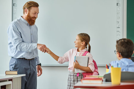 A male teacher in a vibrant classroom shakes hands with a young girl, fostering connection and respect between them.の写真素材