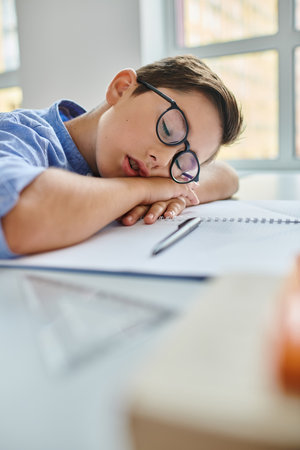 boy wearing glasses peacefully sleeps on a desk in a bright, lively classroom settingの写真素材