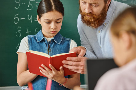 A man teacher reading a book to a young girl with an interested expression in a lively classroom settingの写真素材