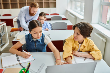 A group of children sit at desks in a bright classroom, listening attentively to a man teacher instructing them.の写真素材