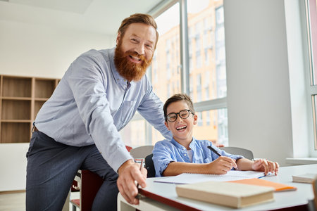 A man standing next to a boy at a desk, engaged in a learning activity in a vibrant classroom setting.の写真素材