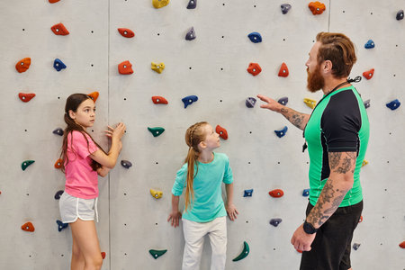 A male teacher is instructing two young girls standing in front of a climbing wall in a bright, lively classroom setting.の写真素材