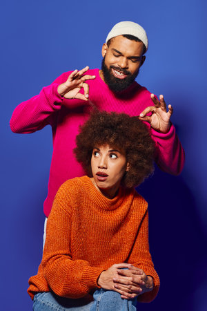 A young African American man and woman, friends, sit closely together in vibrant casual attire against a blue background.の写真素材