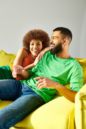 An African American man and woman, dressed vibrantly, share a moment of friendship while sitting on a yellow couch against a grey backdrop.の写真素材