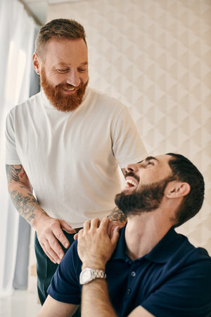 Two men, one with a beard, laughing together in a modern living room. Happy gay couple in casual clothes show love and joy.の写真素材