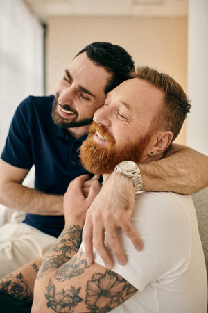 Two men with tattoos embrace affectionately on a comfortable couch in a modern living room, reflecting love and happiness.の写真素材