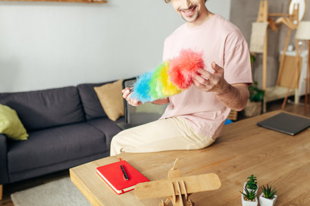A man in cozy homewear sits on a table, holding a vibrant duster.の写真素材