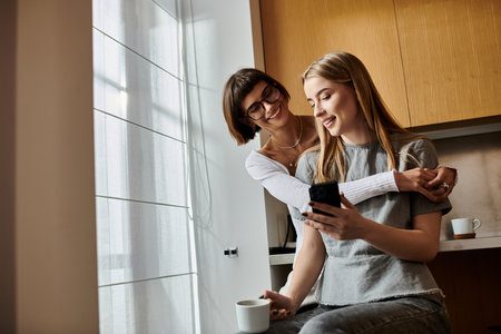 Two young women in a hotel kitchen, sipping coffee and looking at a cellphone with intrigue.の写真素材