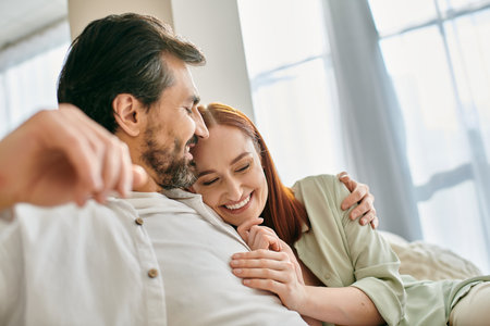 A redhead woman and bearded man share a warm hug while seated on a comfortable couch in their modern apartment.の写真素材