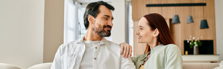 A beautiful adult couple, a redhead woman and a bearded man, sitting on a couch, gazing lovingly at...