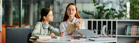 Redheaded woman tutors her teenage daughter, both focused on a laptop during after-school lessons in a modern office setting.の写真素材