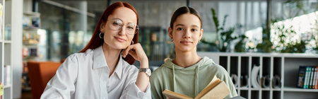 Redhead woman tutors teenage girl in library, both engrossed in bookの写真素材