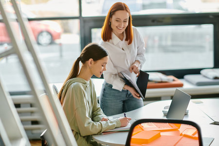 A redhead tutor leads after-school lessons with a teenage girl at a table in an office, utilizing a laptop for modern education.の写真素材