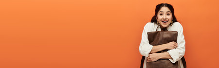 Young indian woman holding brown bag in front of bold orange backdrop.