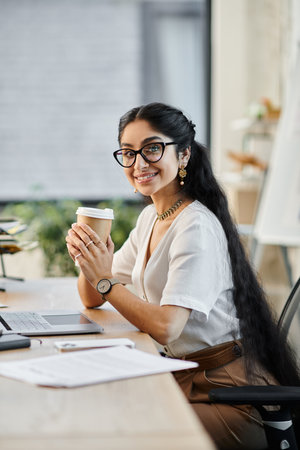 A young indian woman in glasses enjoys a cup of coffee at her desk.の写真素材