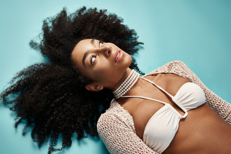 African American woman in trendy bikini lounging on vibrant blue backdrop.
