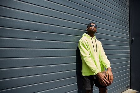 Stylish African American man leans against a wall in a yellow hoodie.