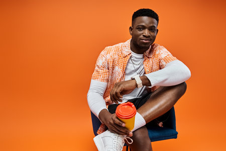Young African American man in chic attire sits, holding coffee.