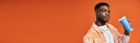 Handsome African American man in stylish attire holding a coffee against an orange background.