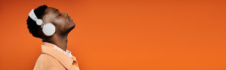 Young African American man in white attire standing in front of vivid orange background with white headphones.の写真素材