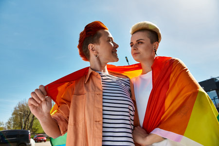 A young lesbian couple proudly stand together outdoors with a rainbow flag, symbolizing love and pride.の写真素材