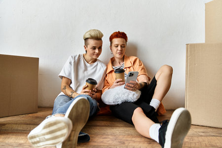A young lesbian couple sits on the ground near boxes at a volunteer center, focused on their cell...
