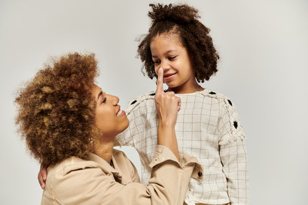 A curly African American mother touching nose of daughter in stylish attire against a grey backdrop.