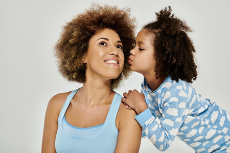 Happy African American mother and daughter wearing pajamas share a tender kiss in front of a white background.の写真素材