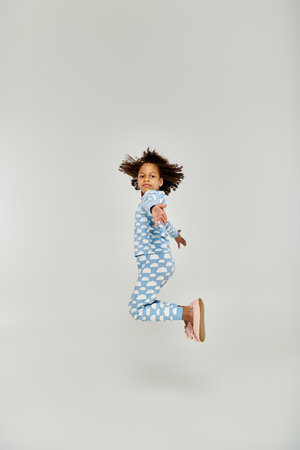 An energetic young girl, dressed in blue pajamas, leaps joyfully in the air on a grey background.の写真素材