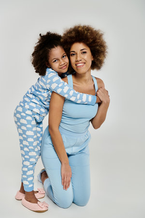 Embracing love and laughter, an African American mother and daughter strike a pose in matching blue pajamas on a grey backdrop.の写真素材