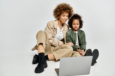Curly African American mother and daughter in stylish clothes sitting on the floor, using a laptop...
