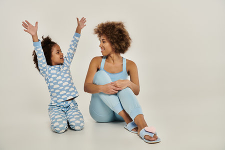 An ecstatic African American mother and daughter in pajamas sitting on the floor, arms raised to the sky in unison.の写真素材