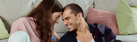 A gay couple, dressed casually, sits close together on a couch, holding glass bong