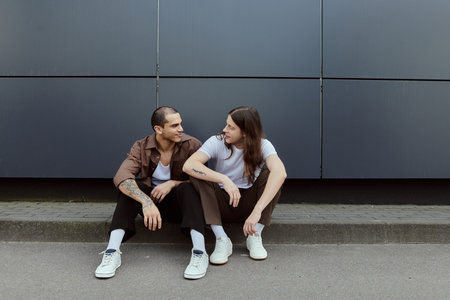 A gay couple in casual attire sits peacefully side by side on the ground outdoors.