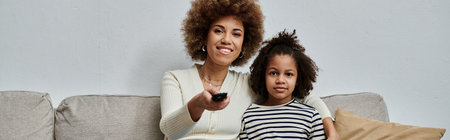 A happy African American mother and daughter sitting on a couch, holding a remote control, enjoying...