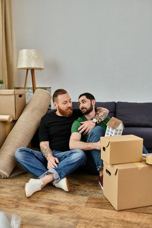 A gay couple relaxes on top of a couch in their new home filled with moving boxes, beginning a new...
