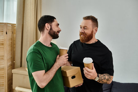 Gay couple standing side by side, holding boxes, ready to move into their new home. Boxes scattered...