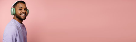 Vibrant young African American man in purple shirt and headphones smiles on pink background.