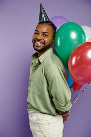 Young African American man in a party hat happily holds colorful balloons on a purple background.の写真素材