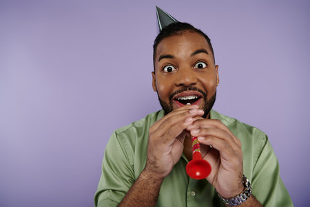 Young African American man joyfully wears a hat while holding a vibrant red party horn against a...