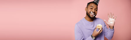 A happy, young African American man with braces, in a purple shirt, savoring a pastry on a pink background.の写真素材